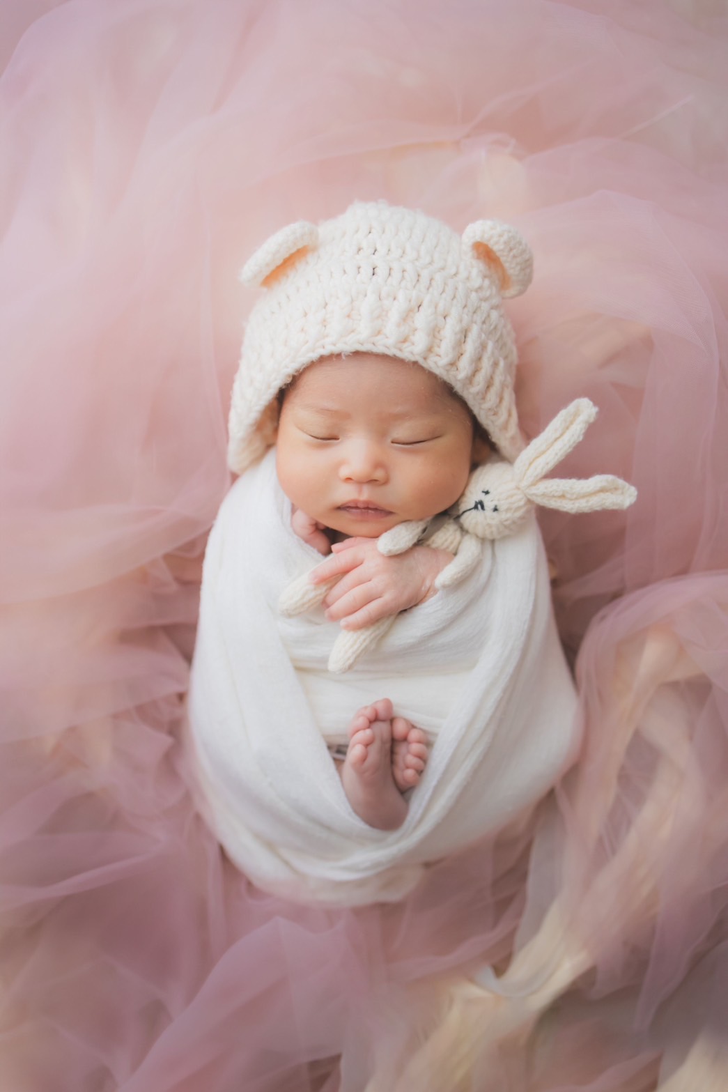 くま耳のニット帽をかぶり、うさぎのぬいぐるみを抱く新生児